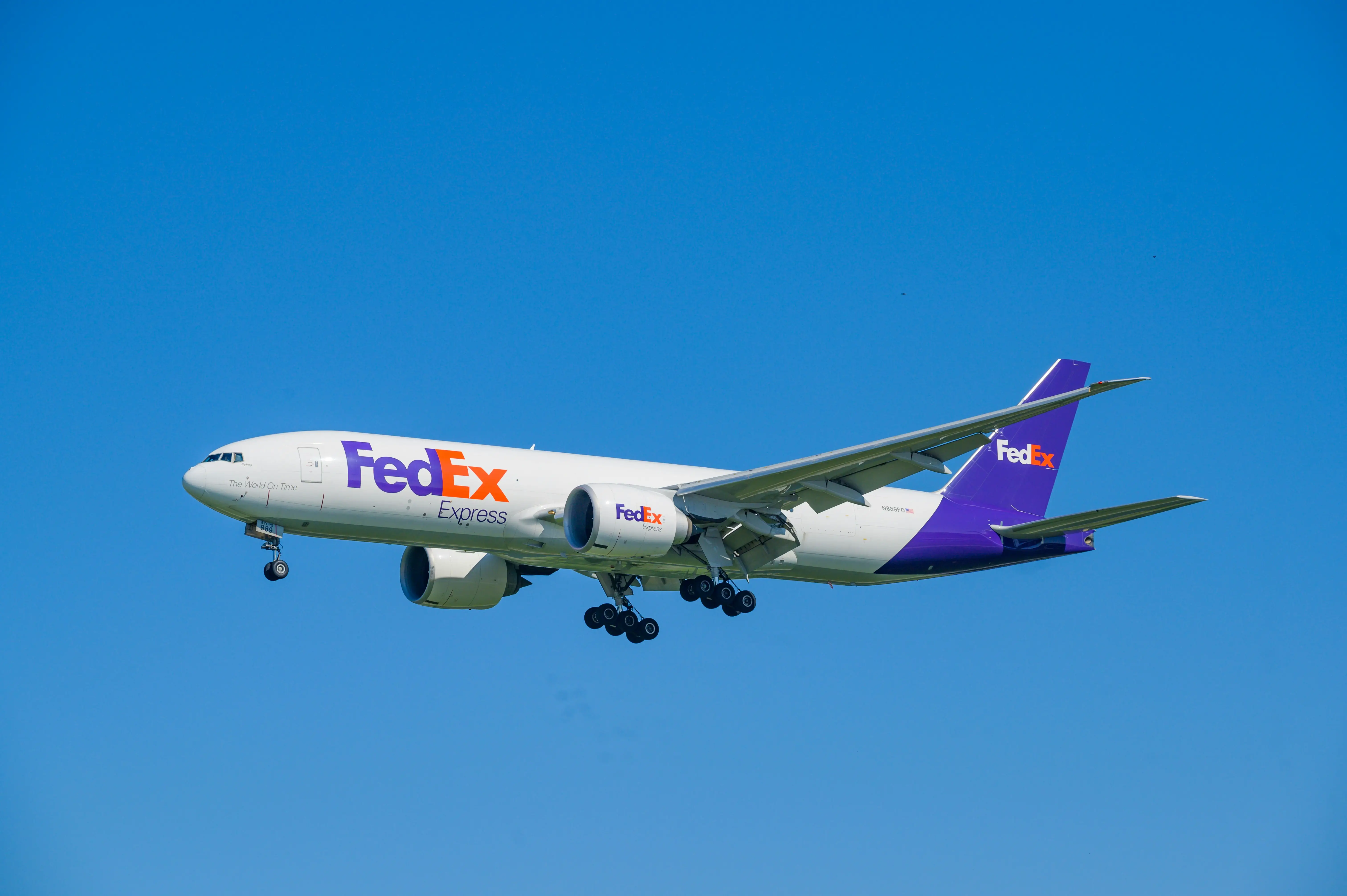 FedEx cargo plane flying with landing gear extended against a clear sky.
