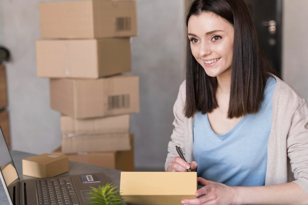 A person at a desk writing on a cardboard box, surrounded by additional packages, suggesting packaging or shipping tasks.