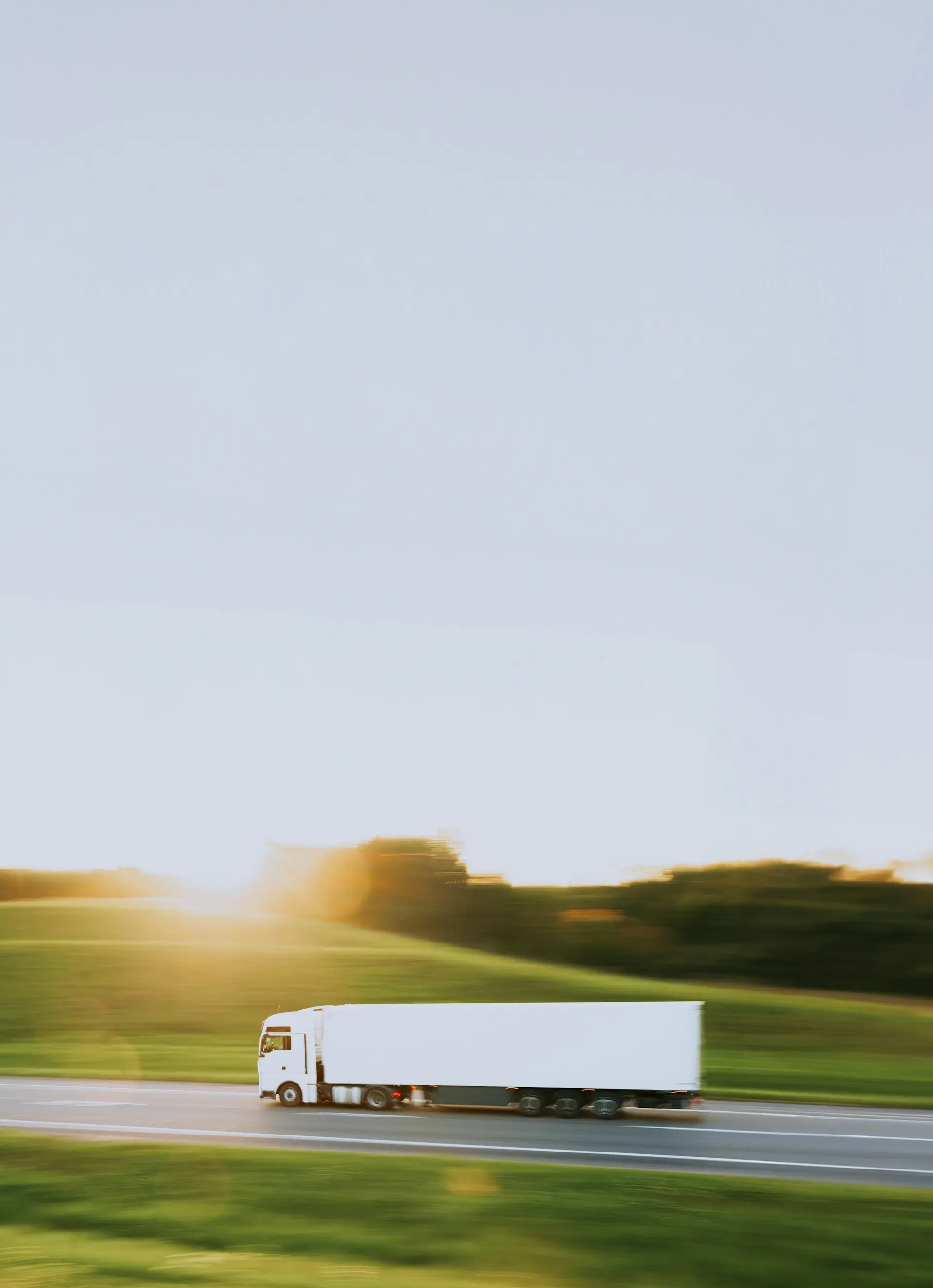 This is a white semi-truck driving on a highway with a blurred background, suggesting motion. The scene includes a green landscape with trees and grass under bright sunlight.