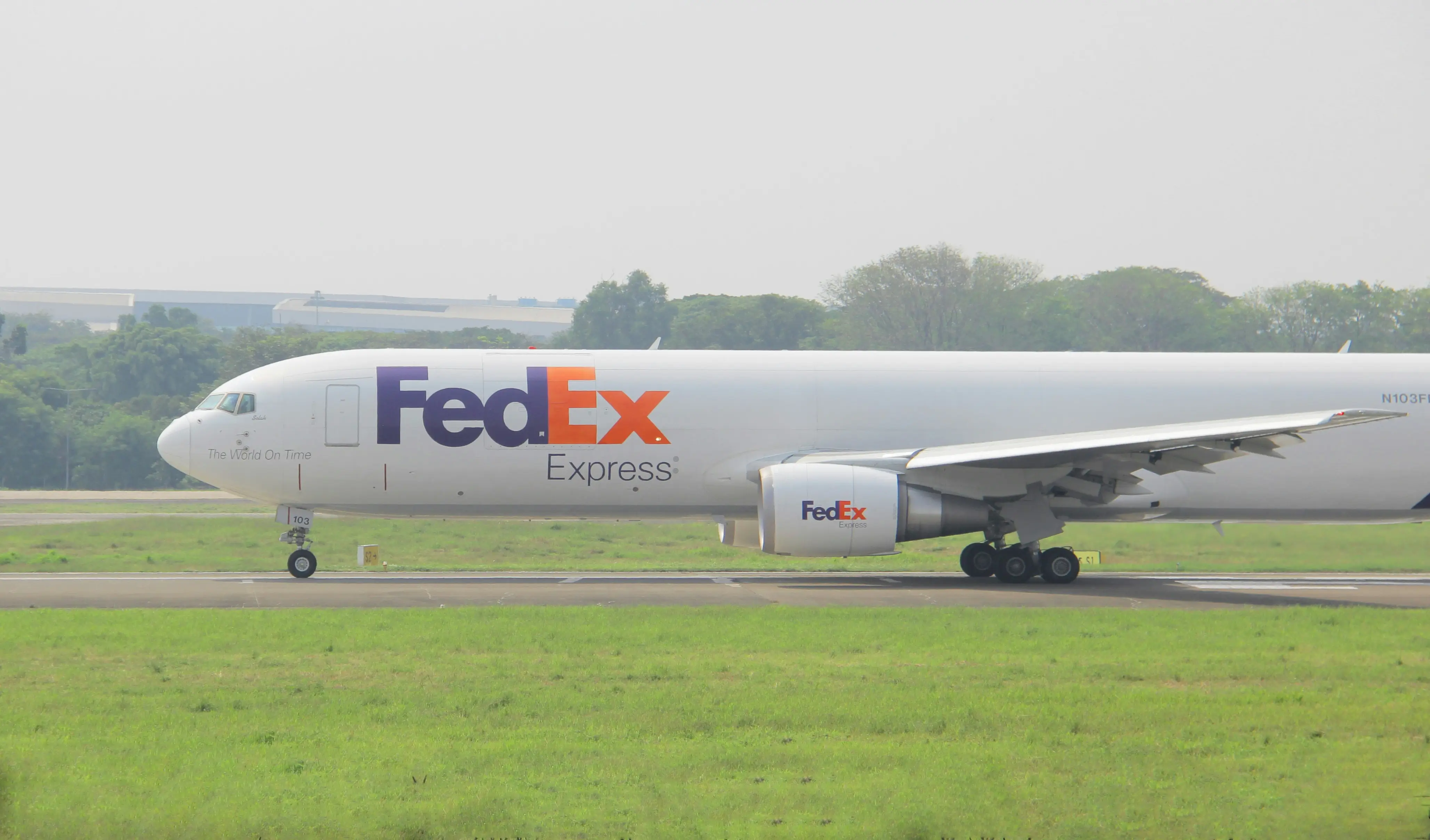 FedEx Express cargo airplane parked on a runway, with greenery and buildings in the background.