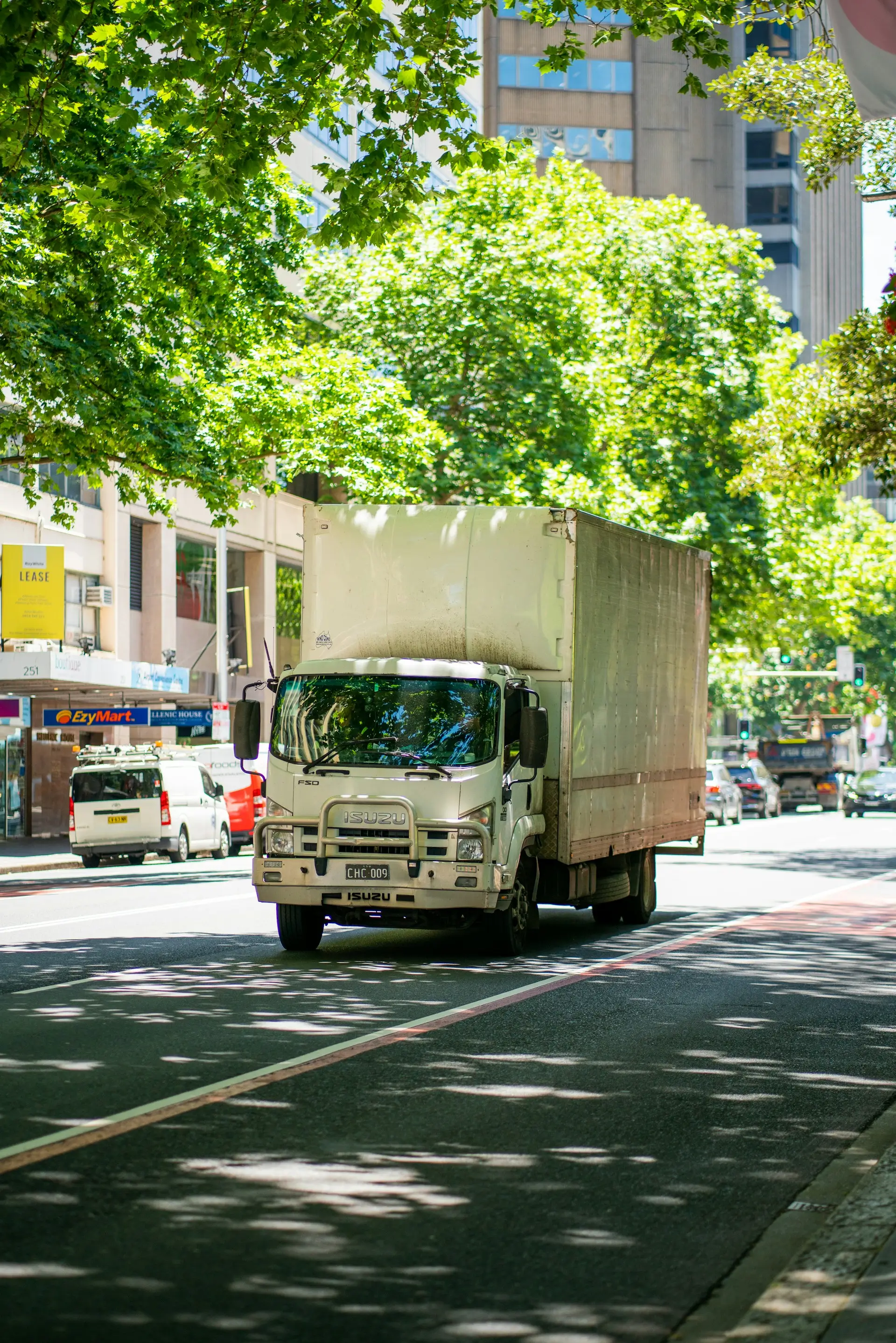 A white Isuzu truck drives down a sunny city street, surrounded by buildings and trees.