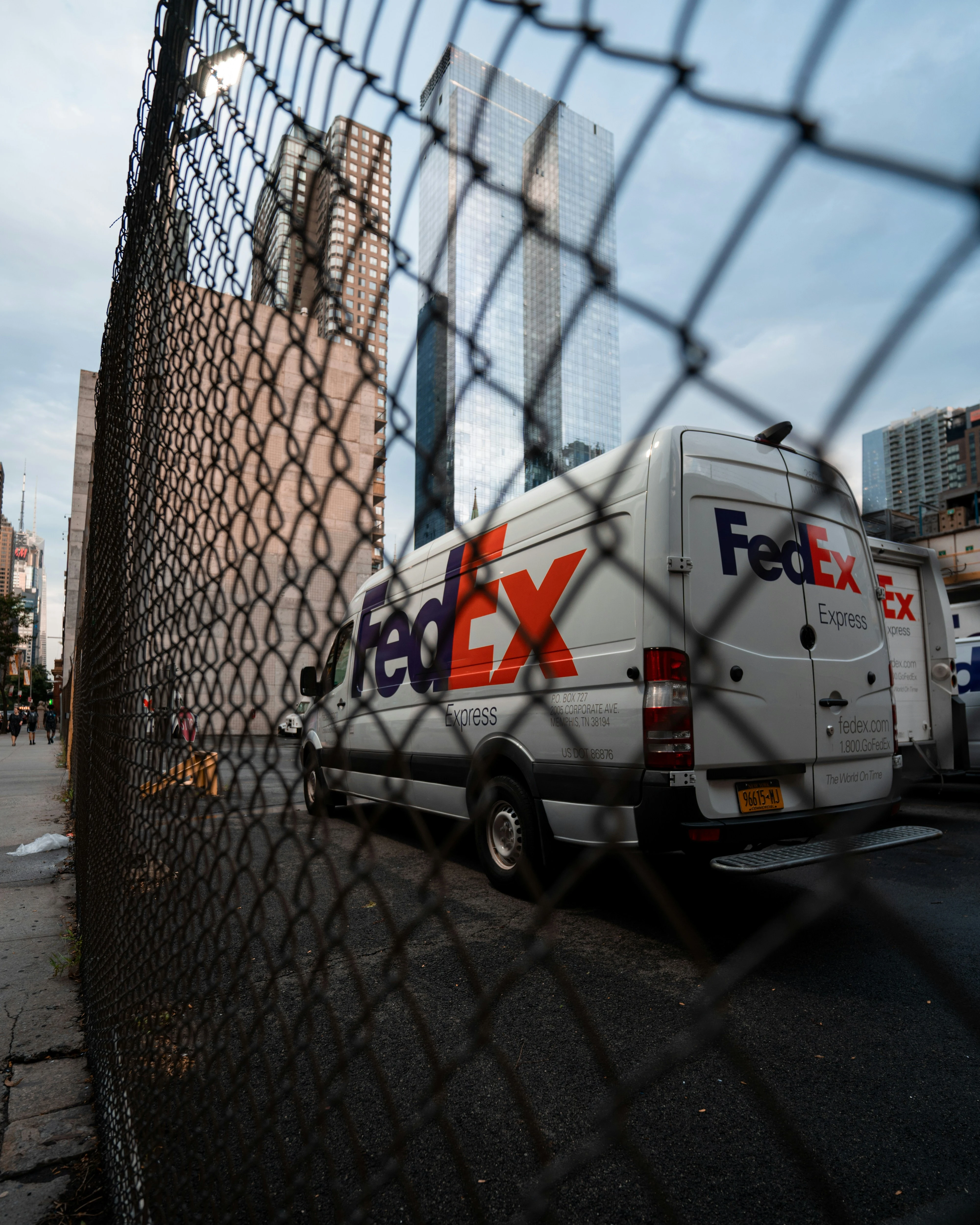 FedEx Express delivery van parked behind a chain-link fence in an urban area.