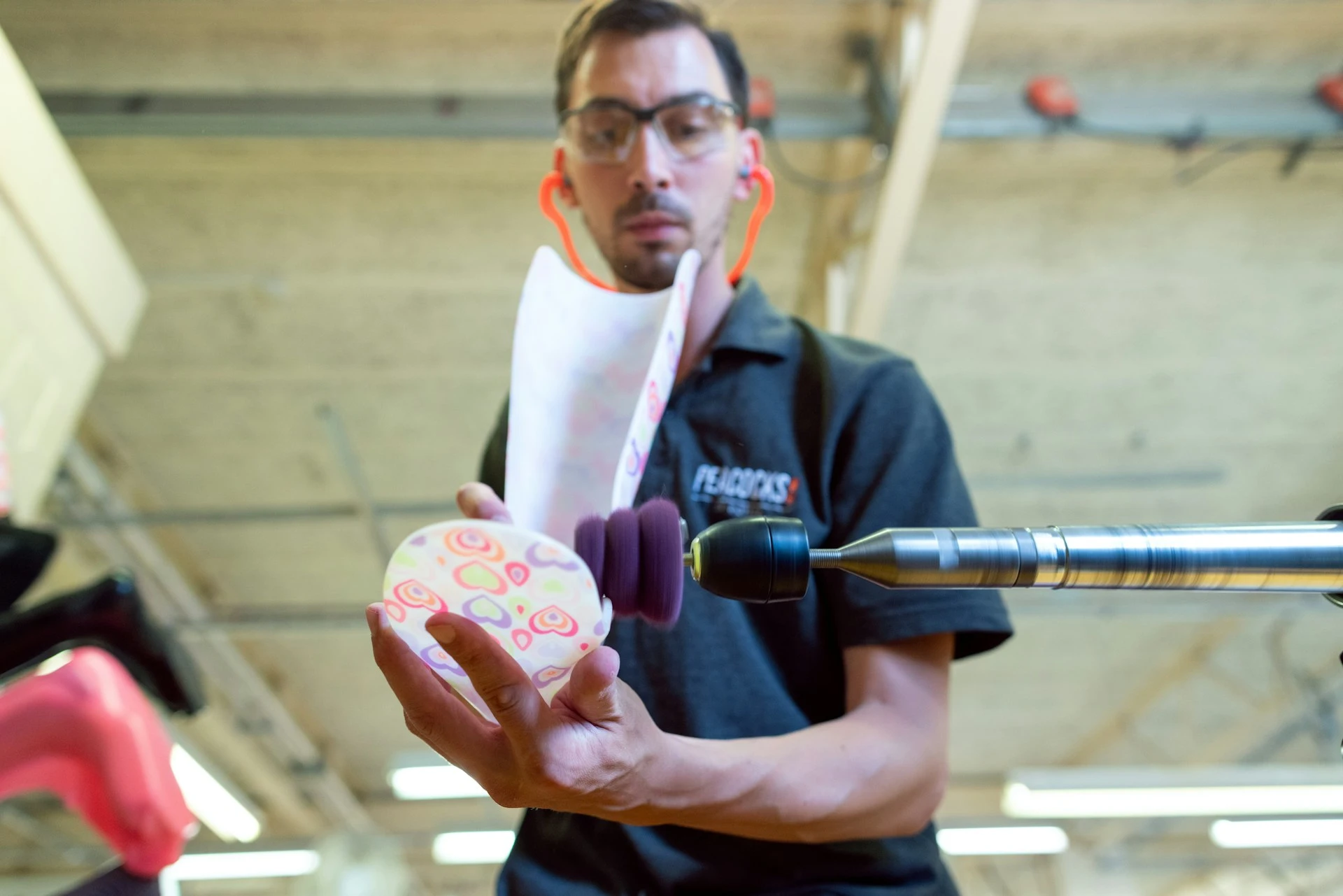 Person in a 'Flexotics' shirt using a drill-like tool on a colorful object in a workshop.