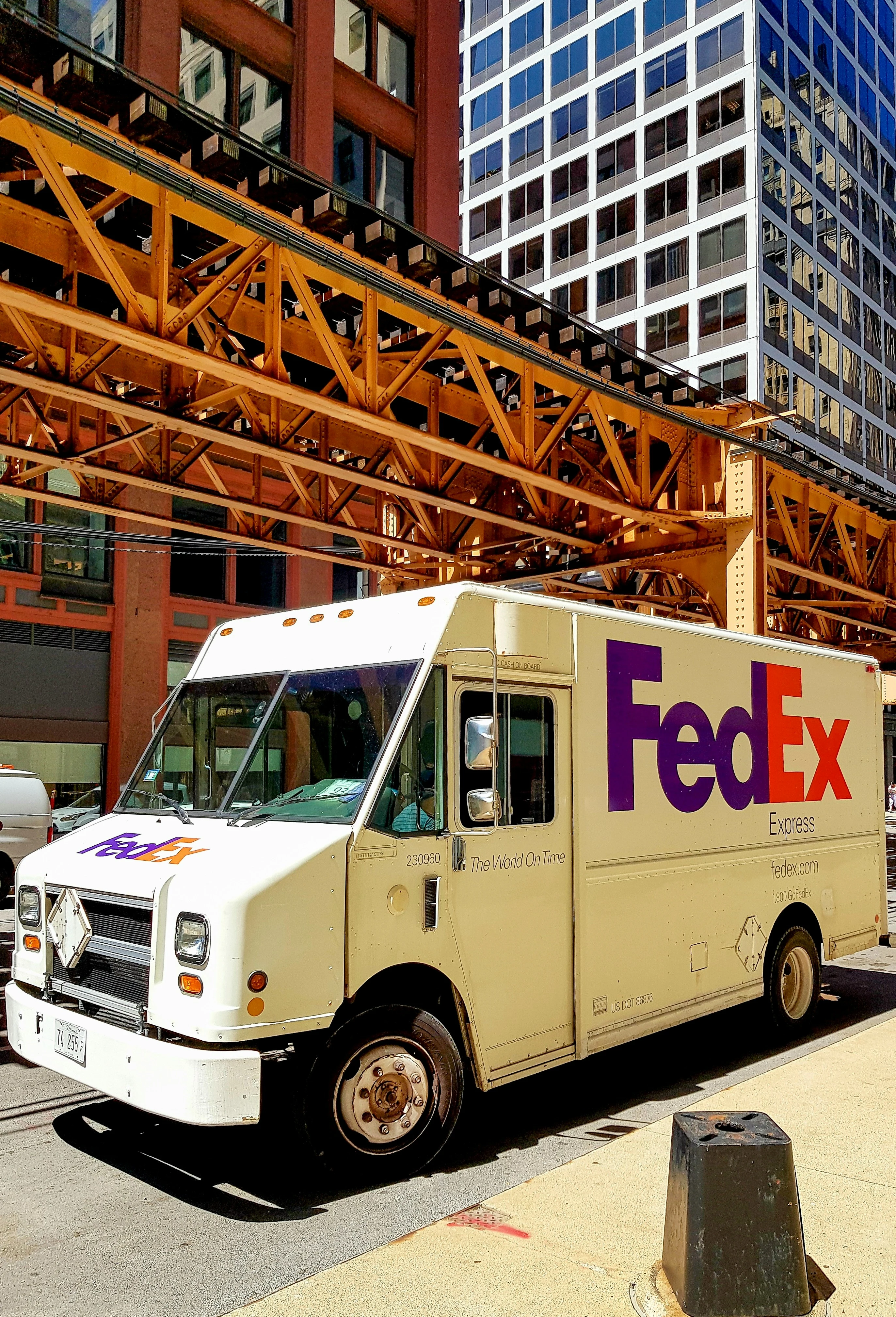 A FedEx delivery truck parked on a city street under an elevated train track.